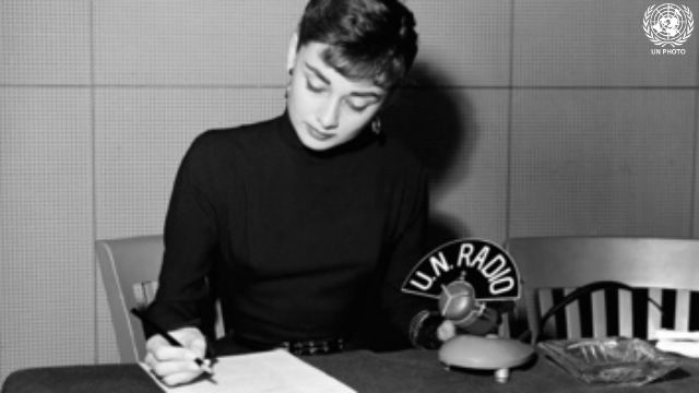 Audrey Hepburn is at a desk with pen and paper and a UN Radio microphone.