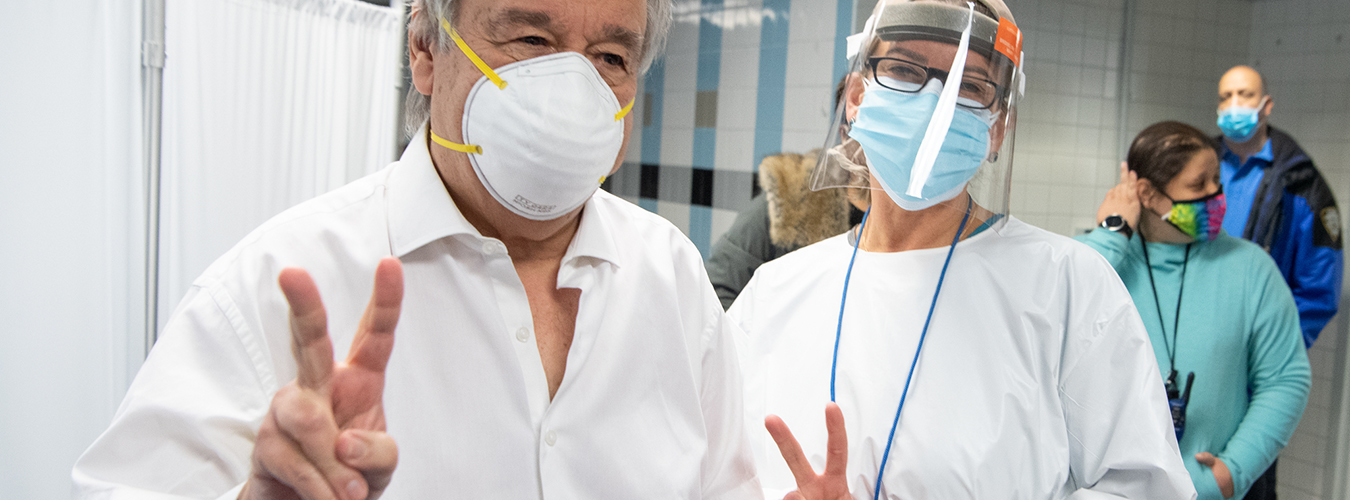 Secretary-General António Guterres and a nurse are wearing masks and both give the victory sign inside a medical setting.
