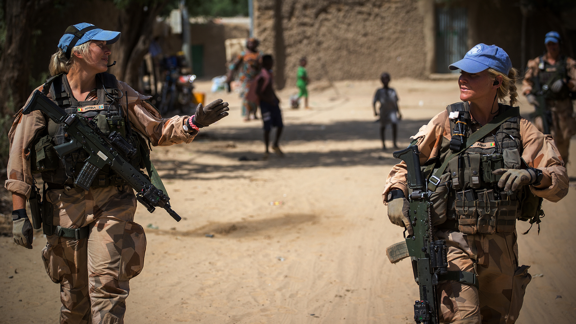 Two female Swedish Marines walk down a street in Mali in 2018 with children in the background.
