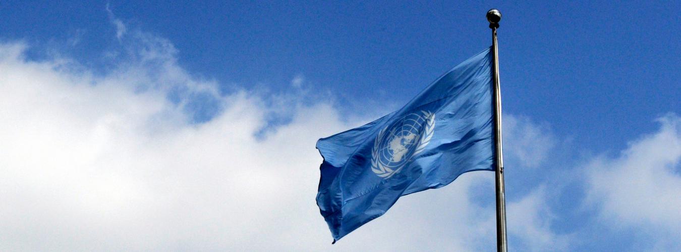 The UN flag flying at UN Headquarters in New York City, with a blue sky in the background.