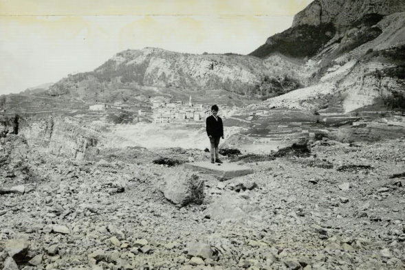 A black and white image of a boy standing in rocky terrain with a town in the background.