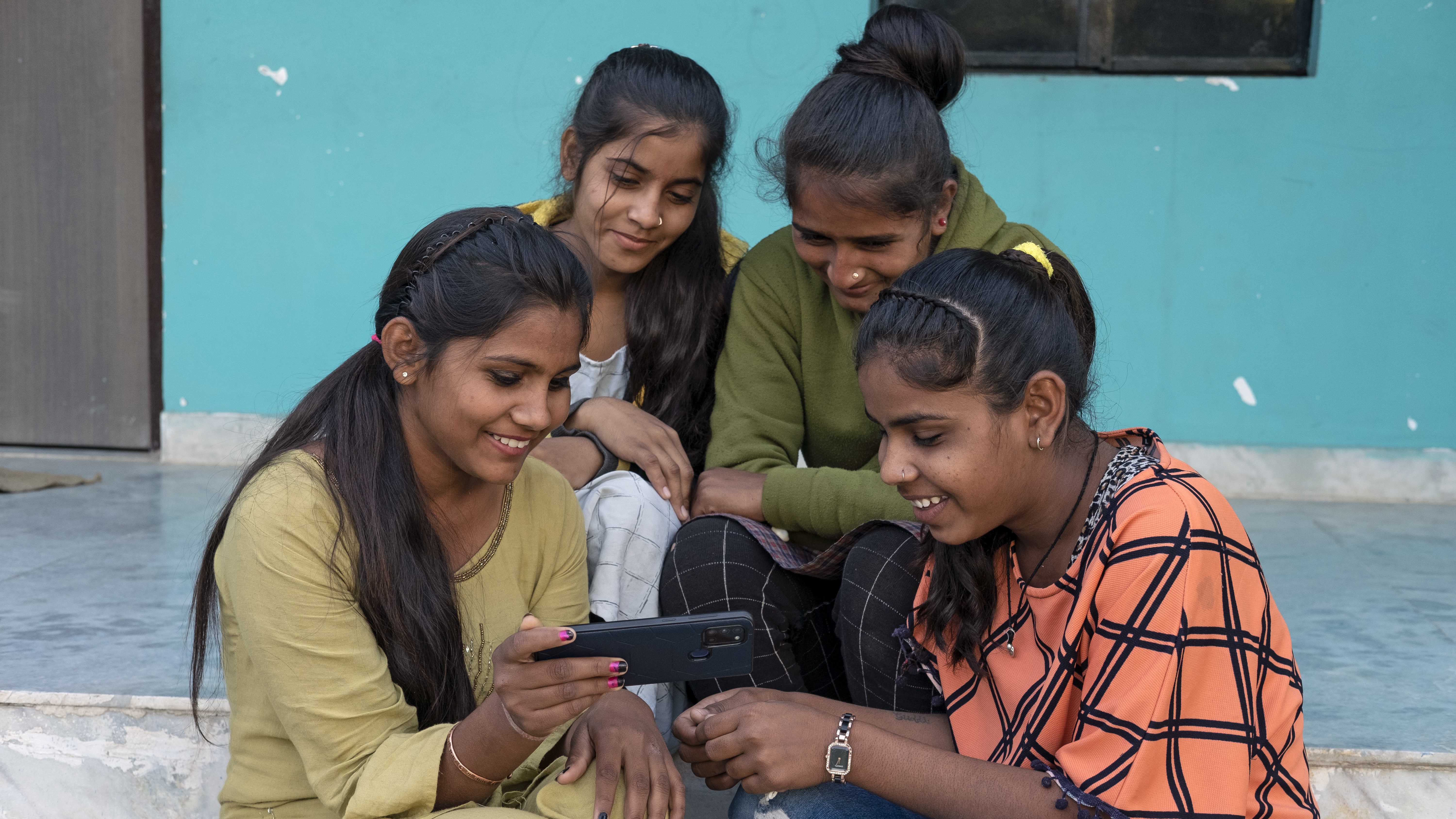 Group of adolescent girls seated on steps look at mobile phone screen.